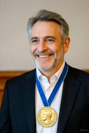 A realistic photograph of Dr. Shimon Sakaguchi, the 2025 Nobel Prize winner in Physiology or Medicine, smiling confidently while wearing a dark suit, white shirt, and the prestigious gold Nobel medal on a blue ribbon around his neck, against a neutral background.