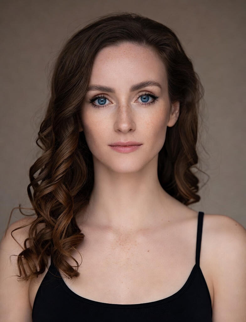 Body Shaming:A young woman with wavy brown hair and subtle makeup stands outdoors in soft sunlight, gazing directly at the camera with a serene expression. Trees blur in the background.
