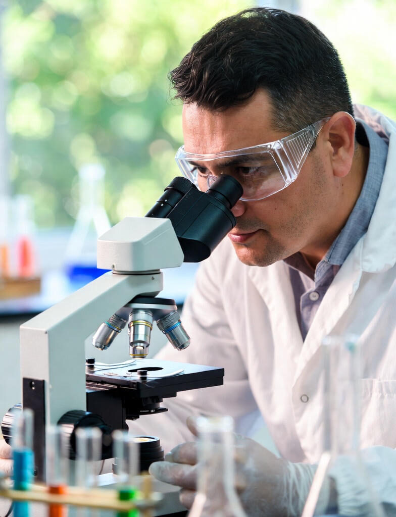 Self-Healing Materials:A male scientist in a white lab coat and safety goggles intently looking through a microscope in a laboratory setting, with colorful test tubes and beakers in the foreground.