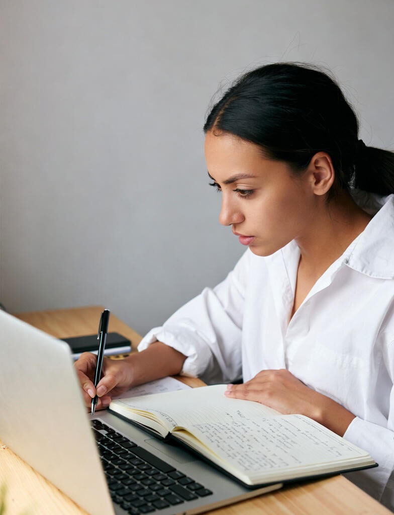 Learn and Earn : Young woman in white shirt focused on studying, taking handwritten notes in a notebook while using a silver laptop on a wooden desk.