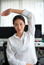 Asian woman in business casual shirt doing an upward arm stretch at her office desk for back pain relief