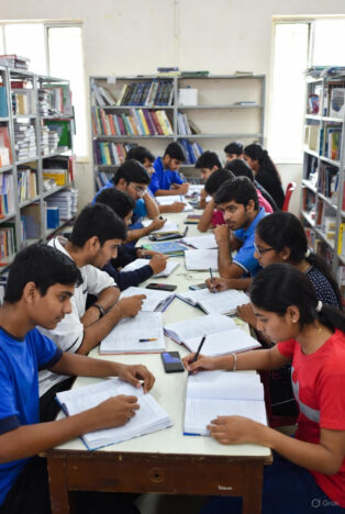 Diverse group of Indian teenagers and young adults studying together at long tables in a bright library or classroom filled with books and notes.