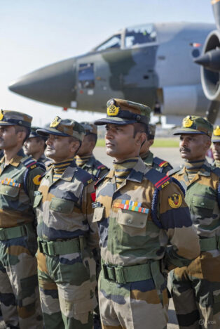 Close-up view of Indian Air Force officers in camouflage parade uniforms with peaked caps and insignia, standing in formation, with a large transport or fighter aircraft prominently in the background.