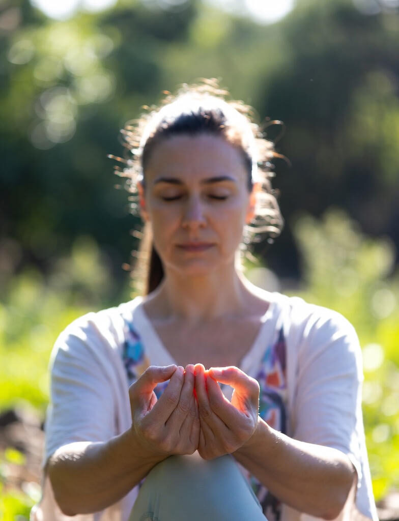 Chakra Activation : A woman in a meditative pose outdoors during morning, with eyes closed, hands in mudra, and soft sunlight illuminating her face against a blurred green background.