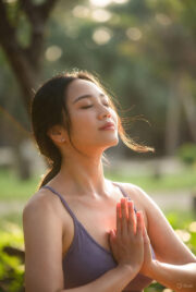 A woman in gentle easy pose (Sukhasana) or modified half-lotus, sitting cross-legged outdoors in a peaceful natural setting, hands in prayer position, eyes closed for calm meditation and fertility support.
