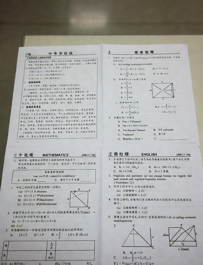Open Gaokao exam papers displaying sections in Chinese Language, Mathematics, English, and Physics, placed on wooden desks in an empty classroom during China
