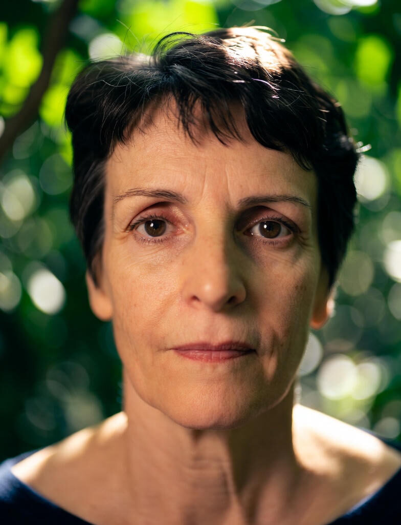 

ALD : Portrait of a middle-aged woman with short dark hair, looking directly at the camera, soft natural light with green bokeh background.