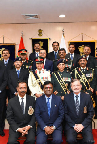 A formal group photo of civil servants, including IPS officers in uniforms and IAS officers in suits, seated and standing in front of the Indian national emblem and flags.