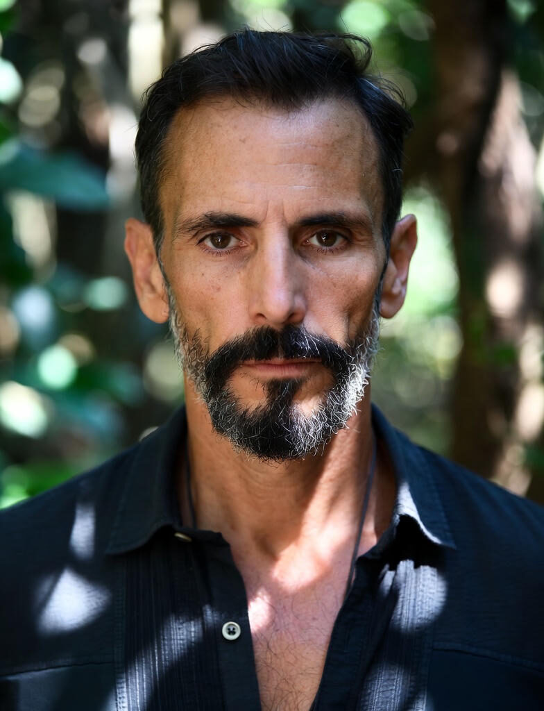 Close-up portrait of a focused, serious mature man with graying beard and dark hair, gazing directly at the camera in a blurred natural forest background with soft bokeh lights.