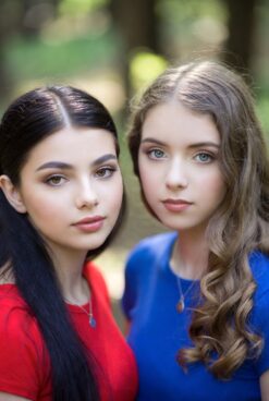 Two happy young women posing closely together in a forest — Indian-origin girl with long black hair in red top and American girl with long wavy brown hair in blue top, both smiling at camera