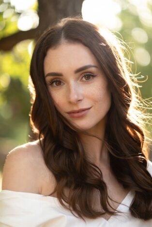 Young woman with green eyes and baby's breath flowers in her curly brown hair, close-up portrait in forest setting representing fertility