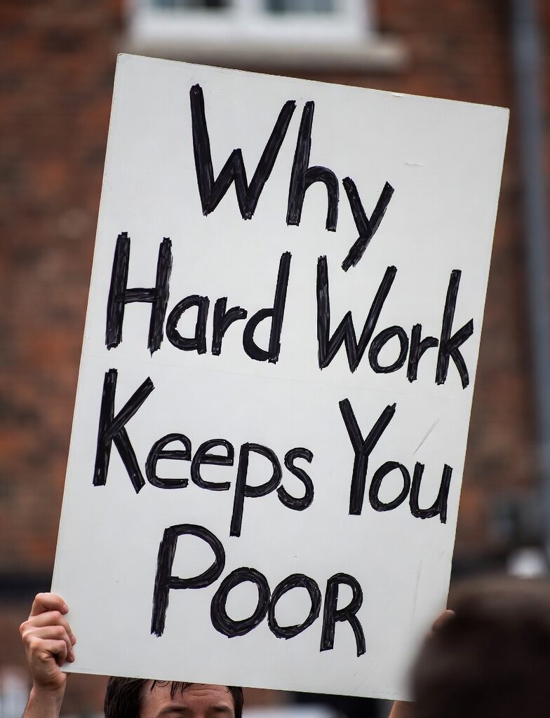 Man holding protest sign reading "Why Hard Work Keeps You Poor" outdoors