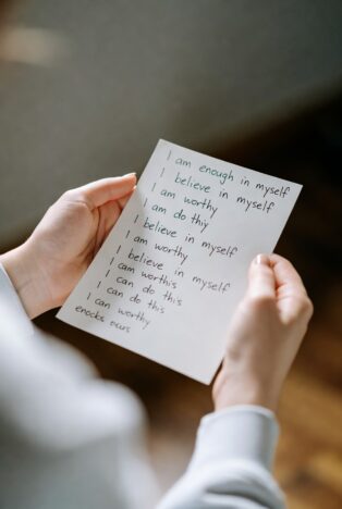 Hand holding a small white card covered in handwritten positive affirmations such as "I am enough", "I believe in myself" and "I can do this"