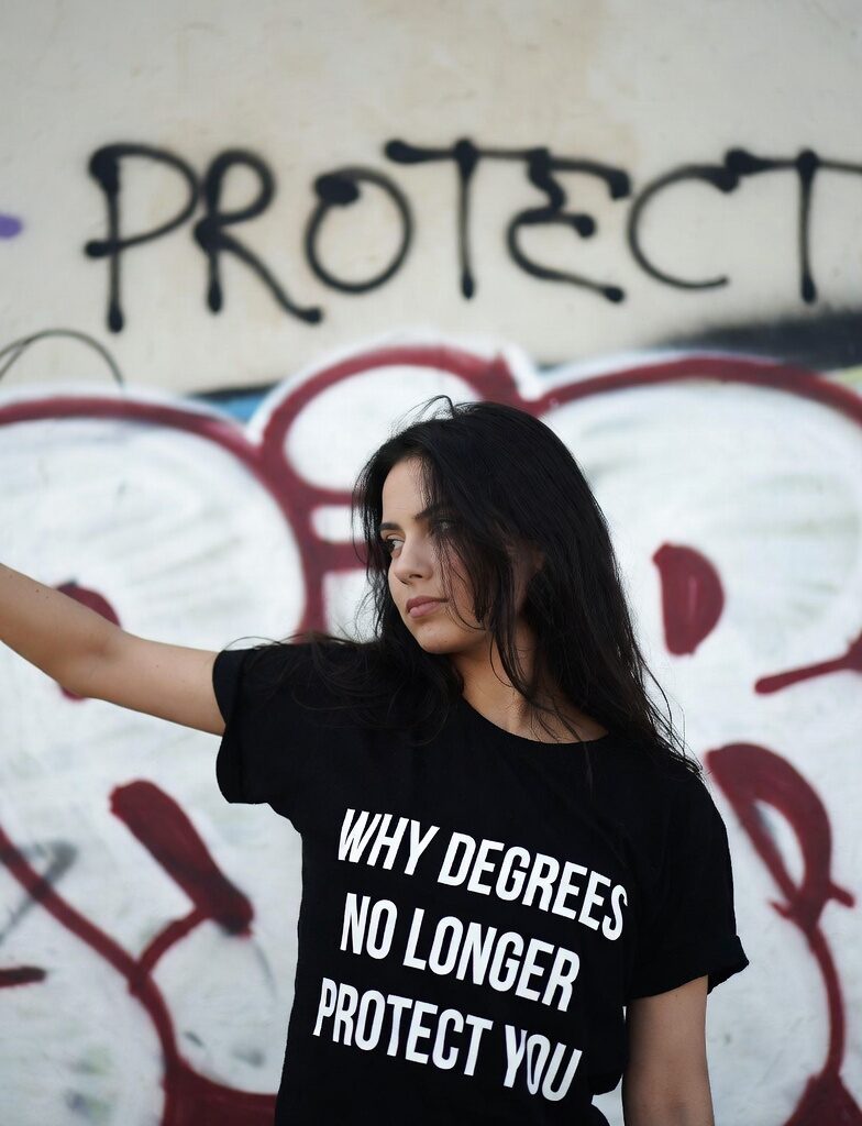 Young woman in black t-shirt with text "WHY DEGREES NO LONGER PROTECT YOU" standing in front of colorful graffiti wall that reads PROTECT
