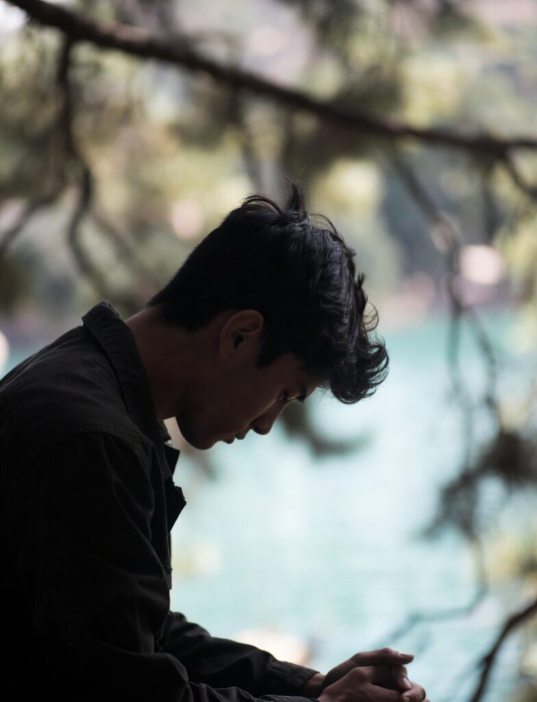 Calm: Young man in dark shirt sitting calmly by a serene turquoise lake, hands clasped, looking downward in quiet reflection under pine trees