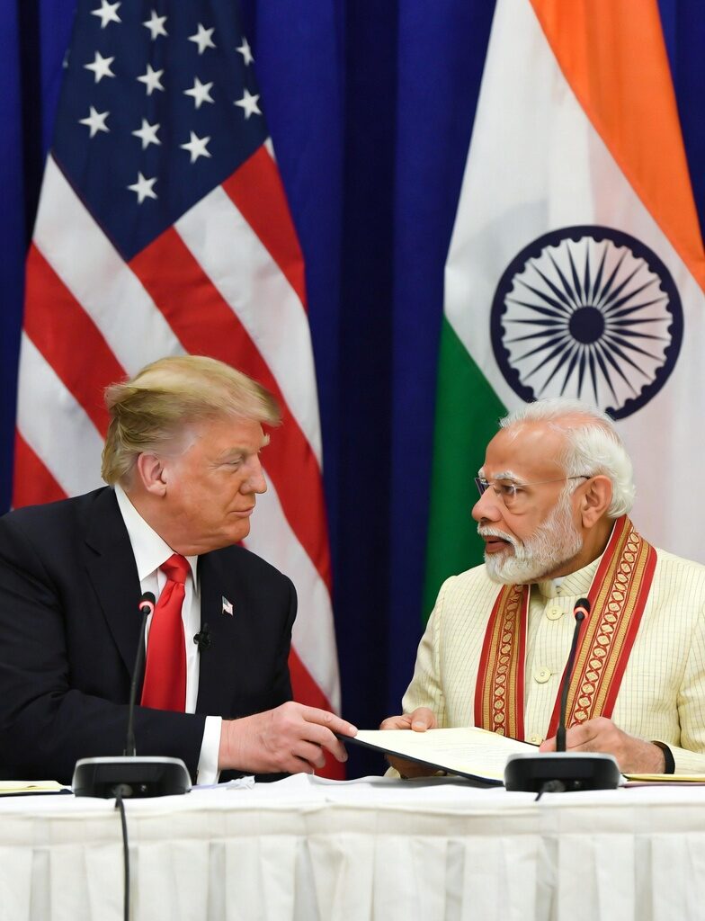 Donald Trump and Narendra Modi seated at a negotiation table, reviewing and signing a bilateral trade agreement between the United States and India, with American and Indian flags in the background.