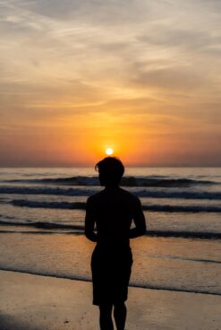 Silhouette of a person standing alone on a beach at sunset, facing the glowing orange sun over calm ocean waves