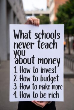 Person holding a sign listing "What schools never teach you about money" with 4 points: 1. How to invest 2. How to budget 3. How to make more 4. How to be rich
