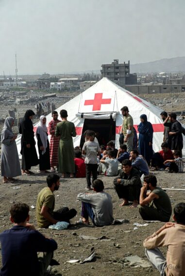 Group of men, women and children sitting on rubble outside a large Red Cross medical tent in a war-damaged city