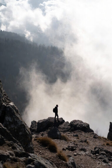 A lone hiker standing on a rocky mountain peak, looking out into a vast sea of clouds and mist, symbolizing the journey of escaping the Minimum Viable Human (MVH) trap.